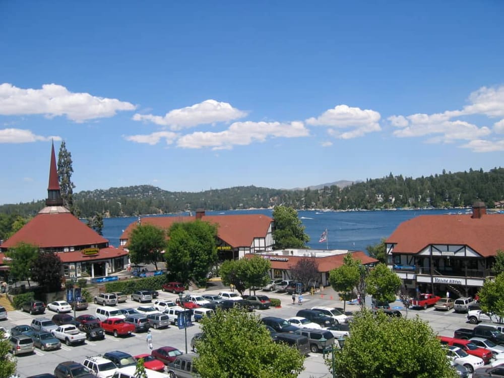 Scenic waterfront shopping area with parking, trees, and buildings near a lake under a blue sky with clouds.
