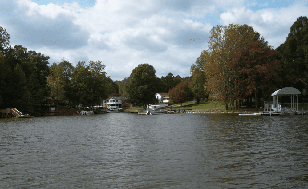 Tranquil lakeside homes with boat docks on peaceful water, scenic fall trees, and cloudy sky.
