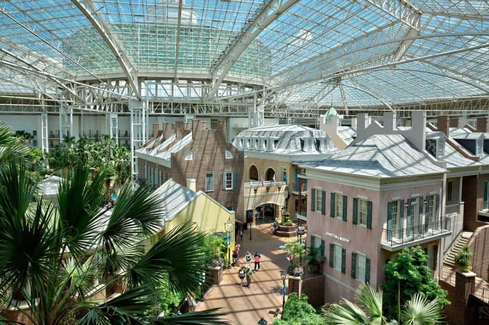 Indoor shopping mall with glass ceiling and European-style storefronts, lush greenery, and shoppers.