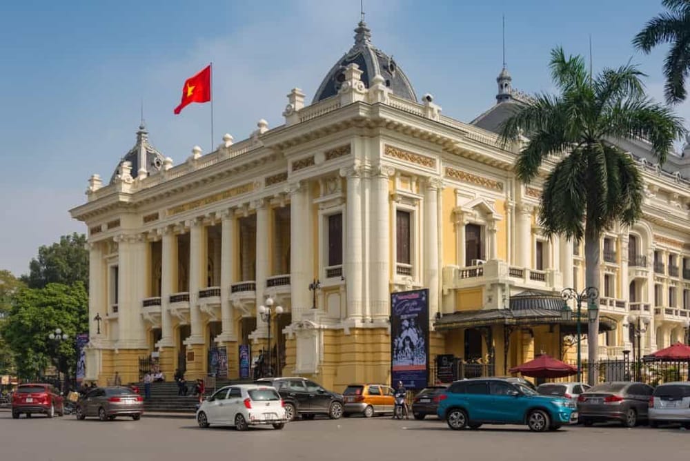Grand French-style historic building with classic architecture in Vietnam, featuring a red flag, palm trees, and busy street.