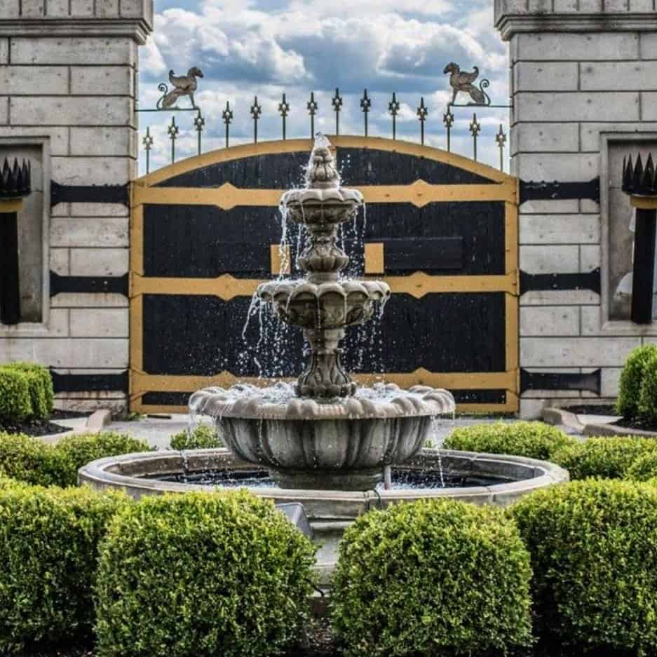 Elegant stone fountain with water cascading, surrounded by manicured bushes in front of a grand estate gate.