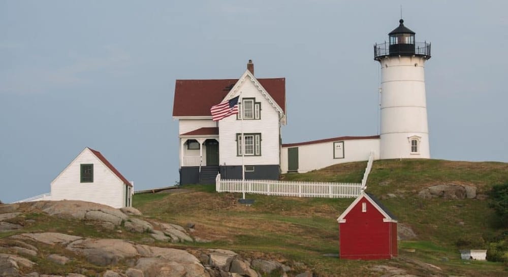 Sunny lighthouse with a historic house and outbuildings on rocky coast, scenic maritime landmark.