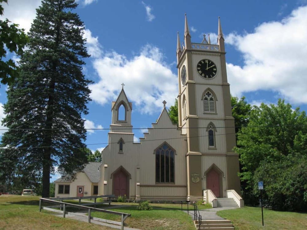 Charming historic church with tall steeple, surrounded by lush greenery and blue sky in a scenic town setting.