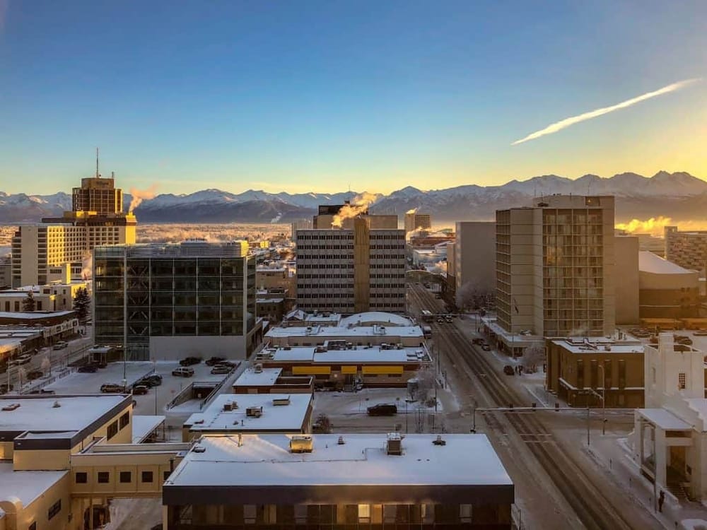 Snow-covered cityscape with mountains in the background at sunrise or sunset, showcasing urban architecture, winter weather, and scenic mountain views.