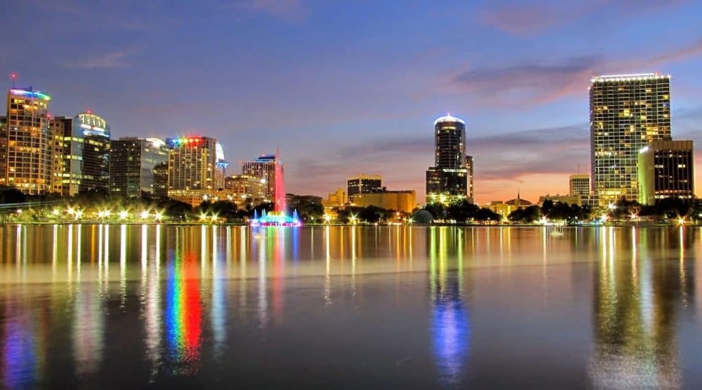 Bright city skyline with lit buildings reflected on water at dusk, showcasing downtown Orlando.