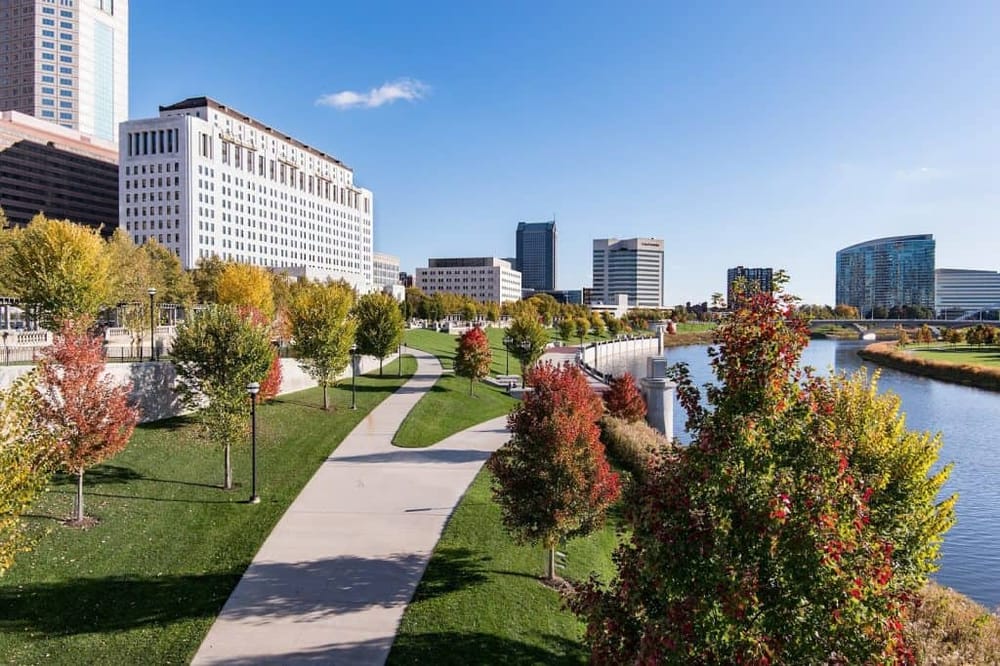 Vibrant city park with walking path by water, autumn trees, and modern downtown skyline.