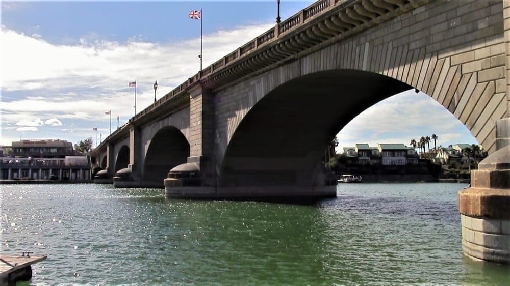 Historic bridge over water with residential area in background, sunny sky, and flags on top.