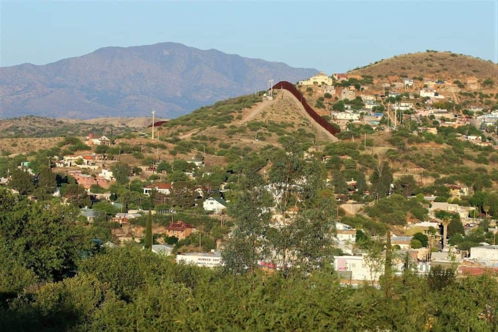 Colorful hillside cityscape with the iconic US-Mexico border wall in the foreground, set against a mountainous backdrop.