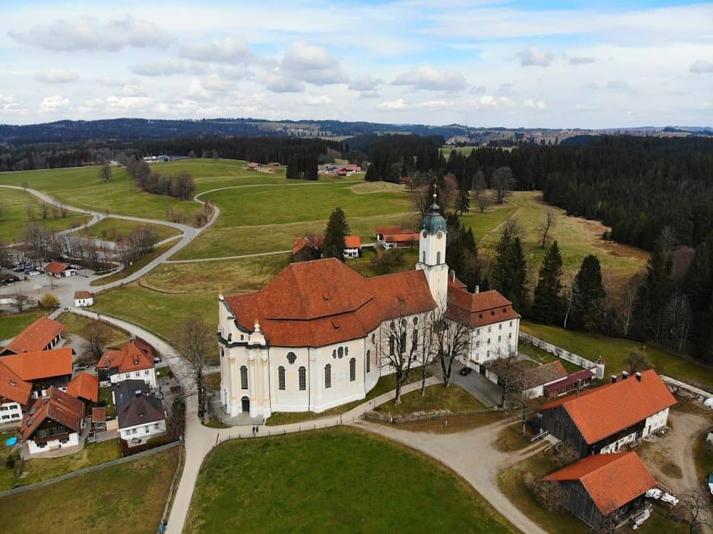 Historic Bavarian church in scenic countryside with rolling green hills and traditional farm buildings.