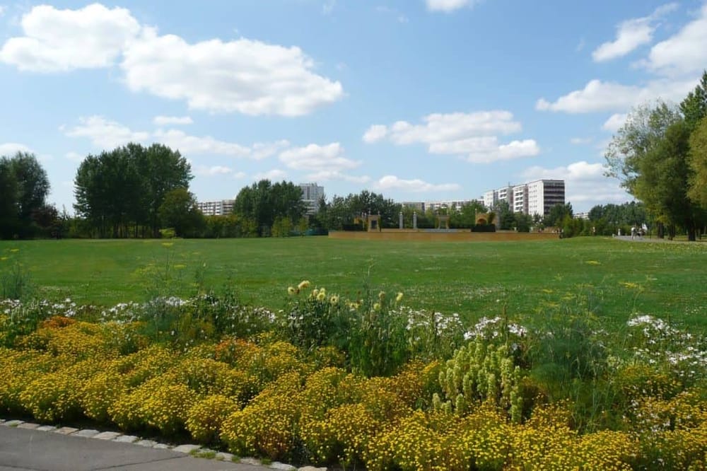 Vibrant city park with flower beds, green lawns, and modern residential buildings under a blue sky.