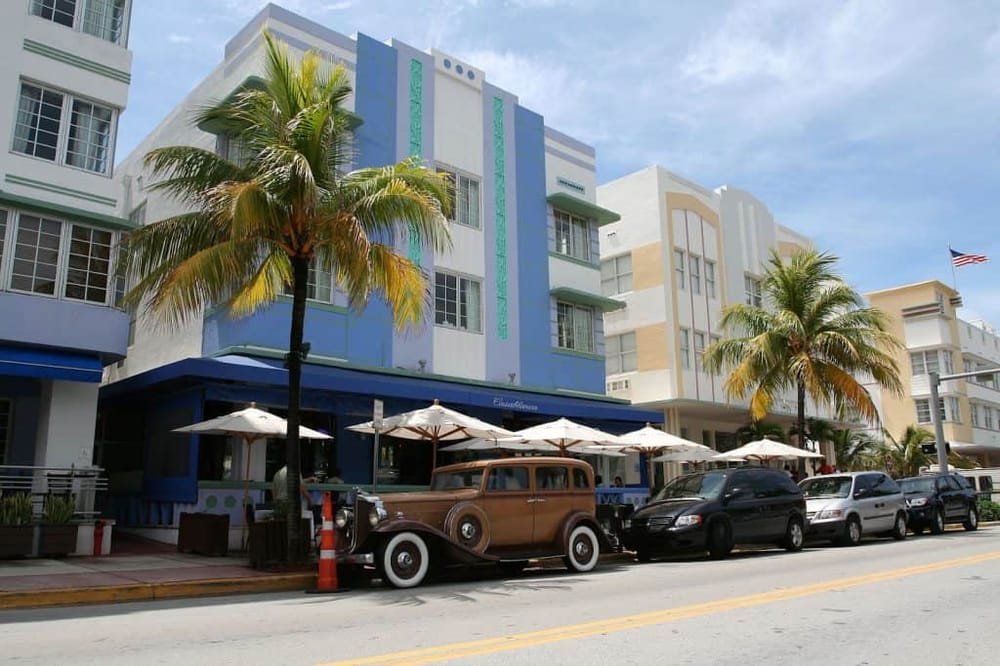 Vintage car parked on Miami Beach street with colorful Art Deco buildings and palm trees.