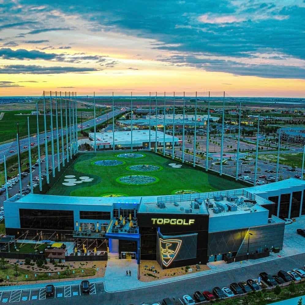 1. Rooftop view of Topgolf venue with golf driving range at sunset, urban shopping and parking lots nearby.