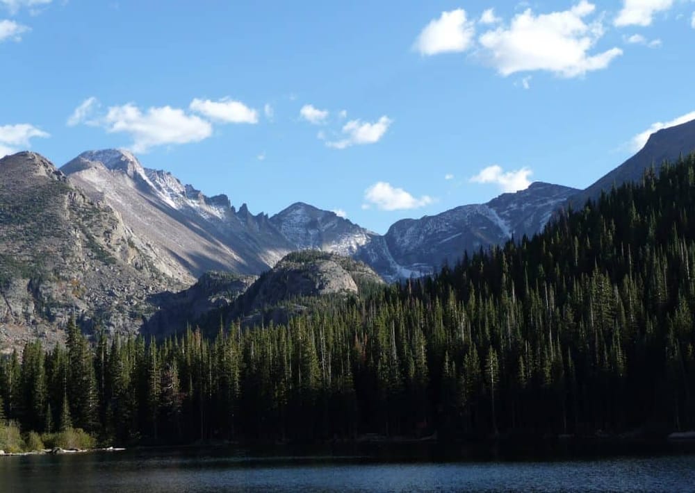 Serene mountain lake with forested shoreline and snow-capped peaks under blue sky.