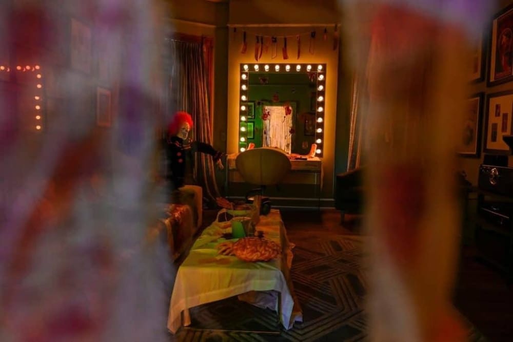 Dimly lit dressing room with mirror surrounded by lights, vintage decor, and Halloween props for a spooky theme.