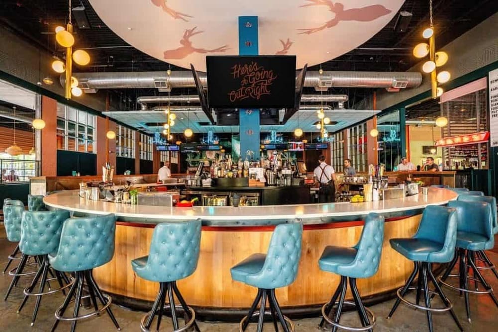 Cozy modern bar with teal tufted chairs, illuminated lighting, and vibrant ceiling art for nightlife enjoyment.