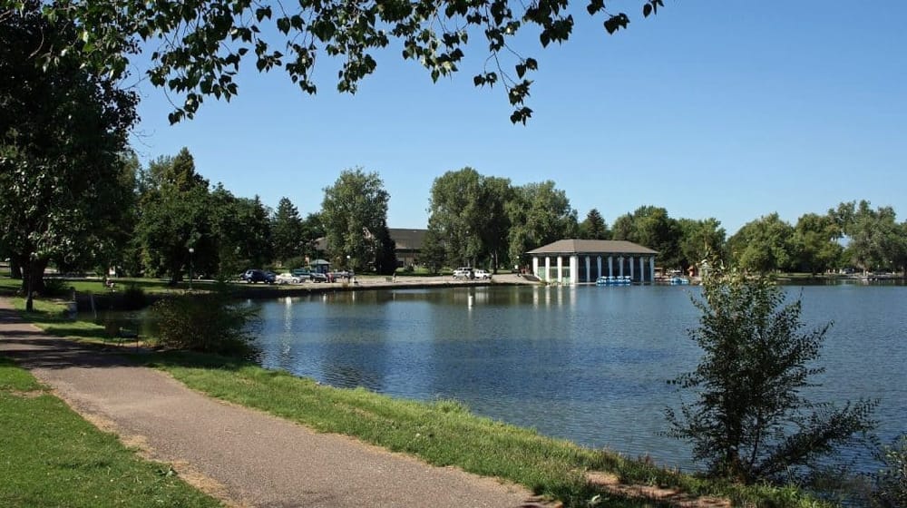 Tranquil lakeside park with walking trail, lush green trees, and a boathouse, perfect for outdoor recreation and community events.