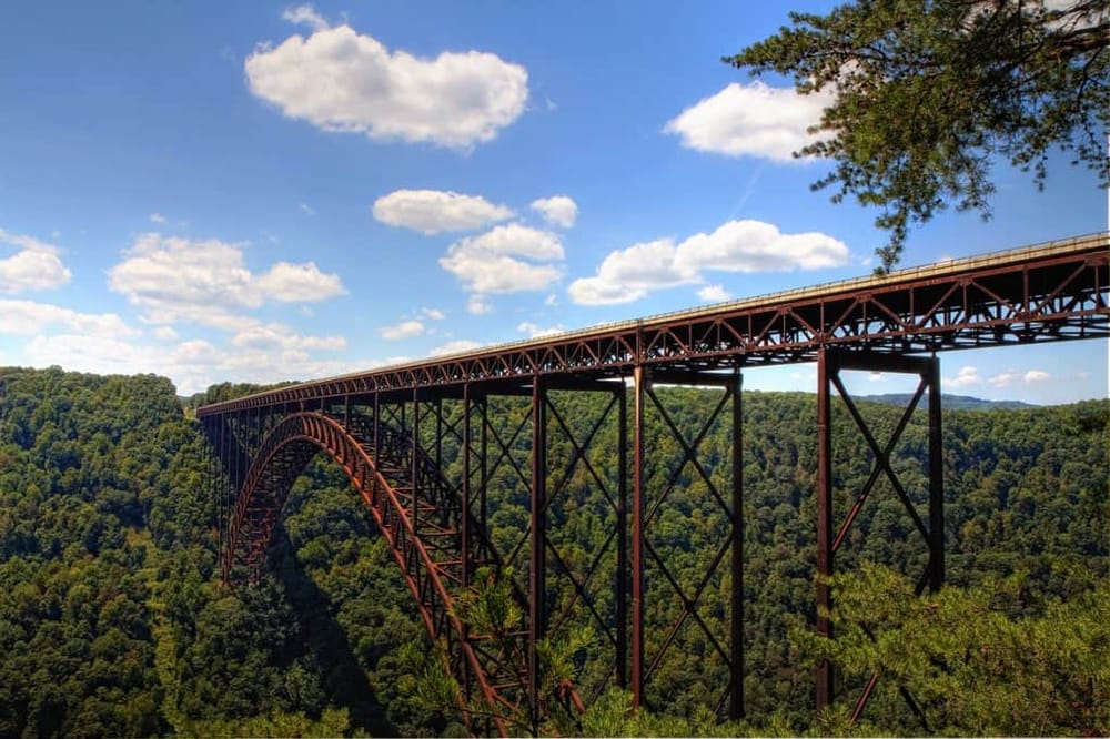 Rustic iron bridge over lush green forest with blue sky and clouds, scenic transportation route.