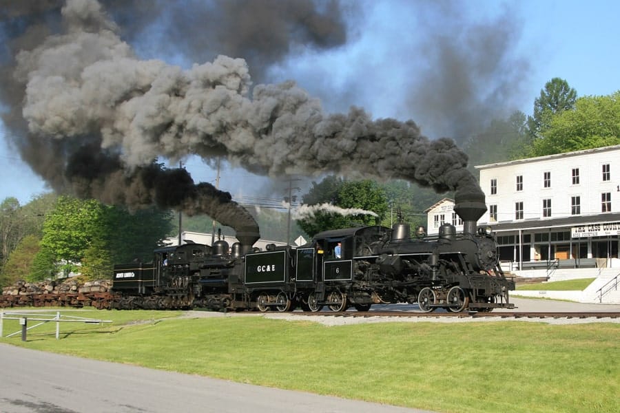 Black steam train emitting thick black smoke on a bright day, historic ride at a scenic location.
