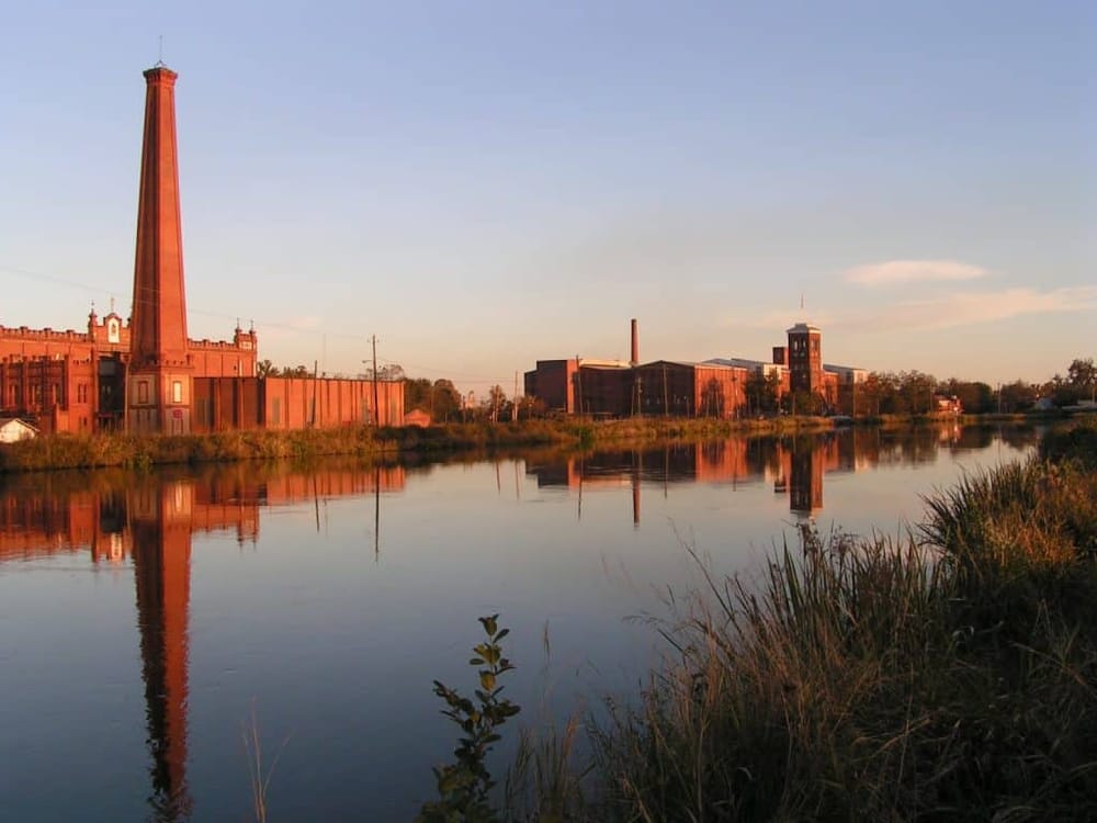 Vintage industrial buildings along a calm river at sunset, reflecting a historic factory setting.