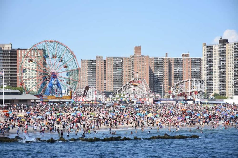 Crowded Coney Island beach and amusement park with Ferris wheel and roller coasters on a sunny day.