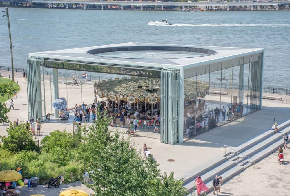 Modern glass pavilion with a carousel inside, located by the waterfront, attracting visitors and families.
