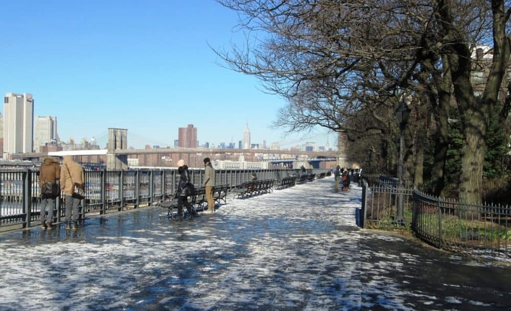 People walking along a NYC waterfront promenade with a view of the Manhattan skyline in winter.
