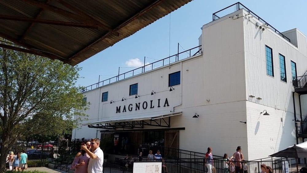 Bright white Magnolia building with black signage, outdoor seating, and people enjoying a sunny day.