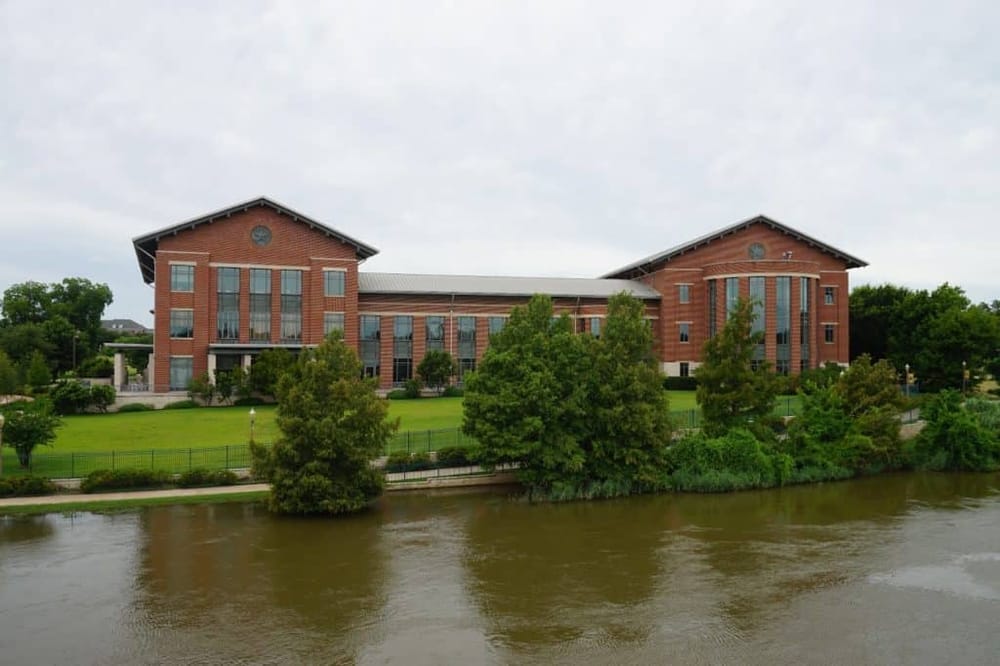 Contemporary brick building with large windows along the river, surrounded by lush greenery and landscaped grounds.