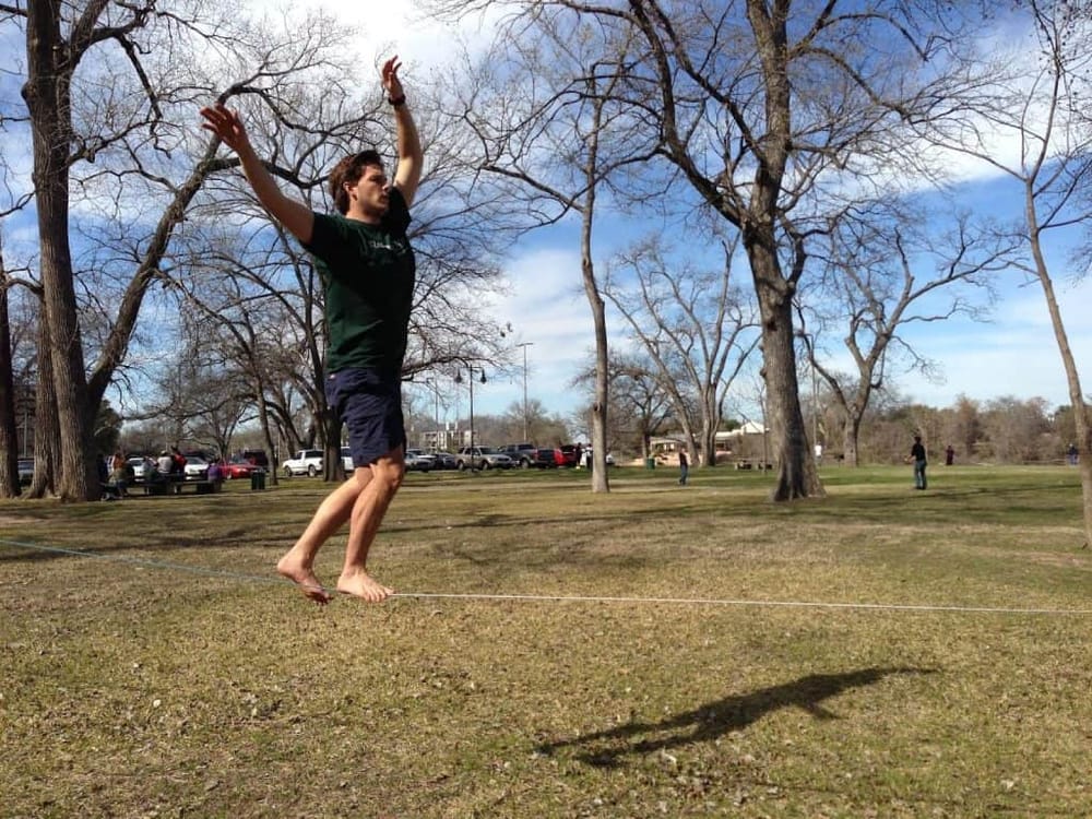 Balanced man walking on slackline in park with trees and blue sky.