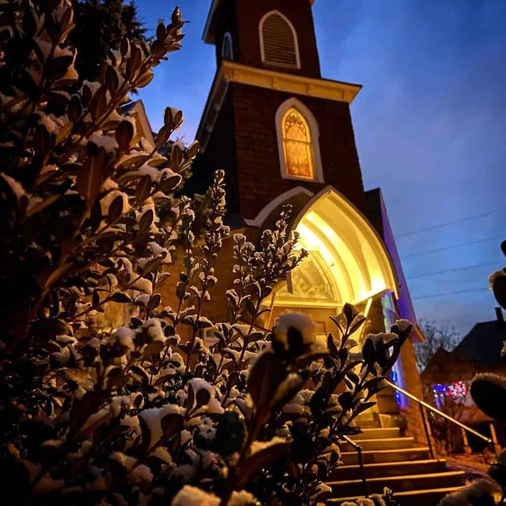 Warm lit church during evening with snow-covered bushes in foreground.