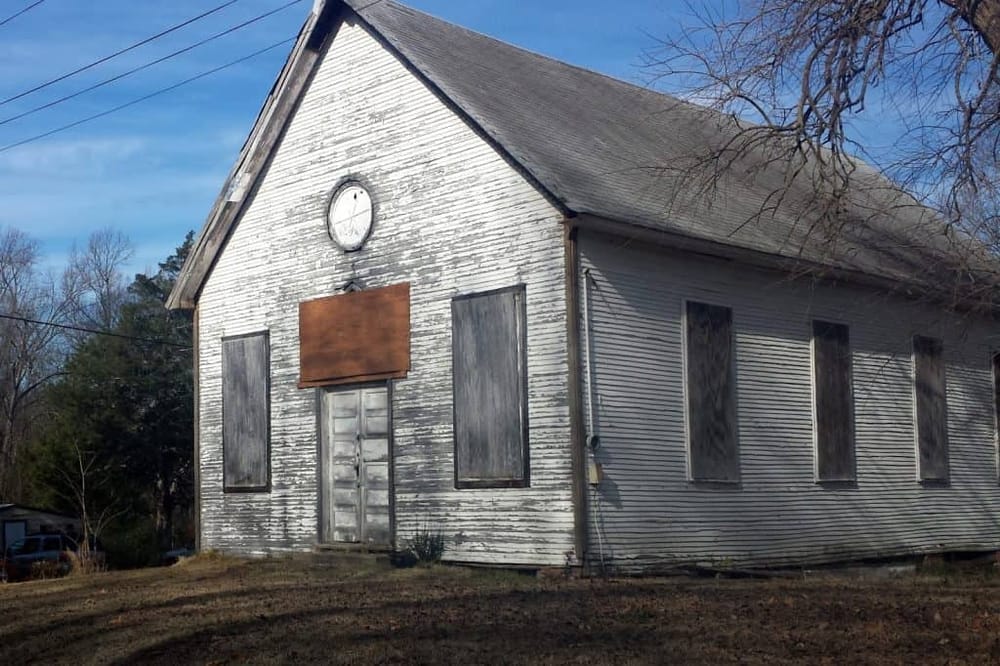 Abandoned old church with weathered white wood siding, boarded windows, and a large clock face on the roof.