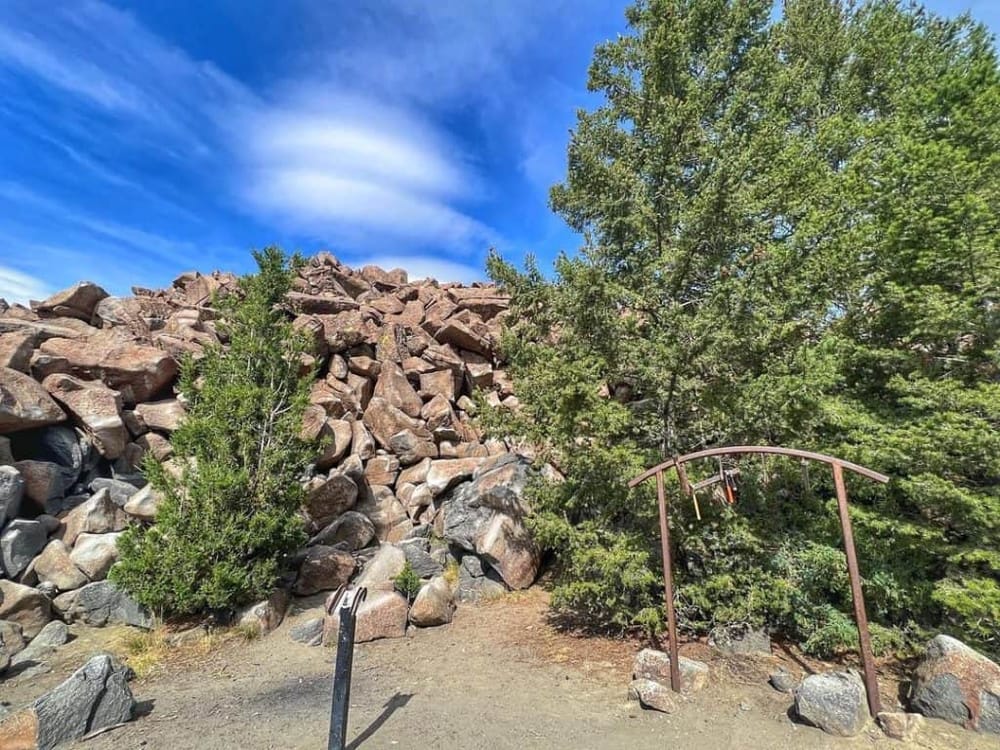Vast outdoor landscape with large rocks and trees under a bright blue sky, featuring a bronze arch with hanging tools.