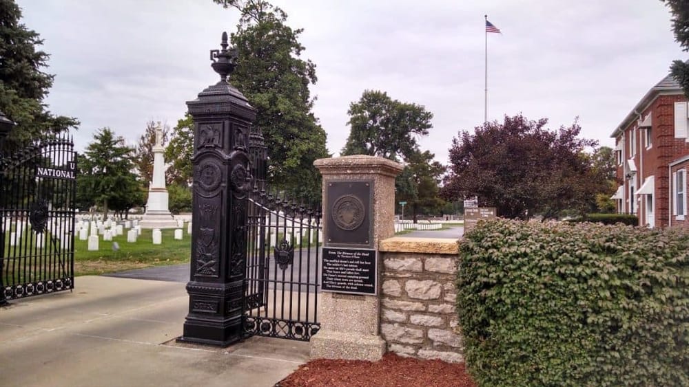 Historic military cemetery entrance with ornate black gate and American flag in background, honoring fallen soldiers.