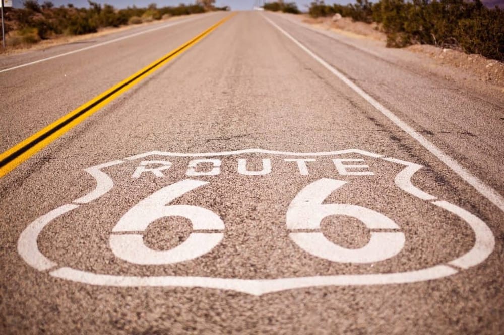 Route 66 road sign painted on highway pavement in desert landscape.