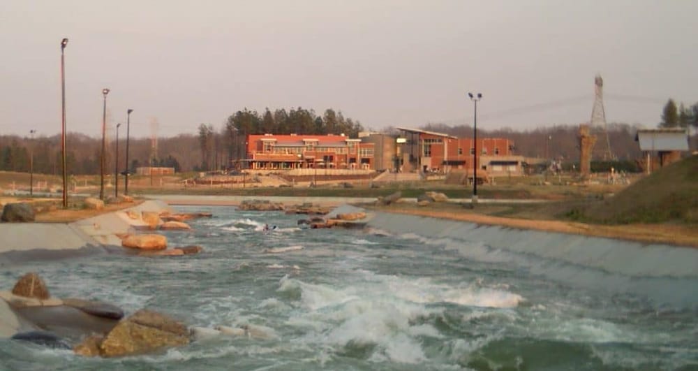 Modern building near water park with rapids and rocks, outdoor lighting and trees in background.
