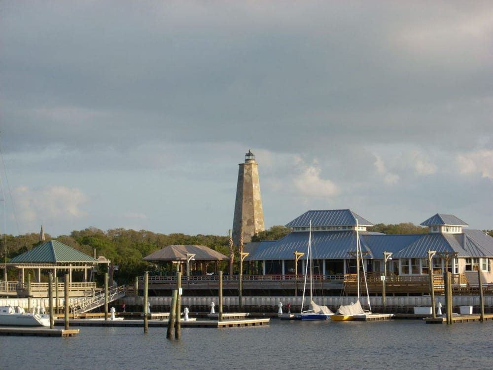 Lighthouse at boat harbor with docks and sailboats, coastal scenery, and waterfront dining area.