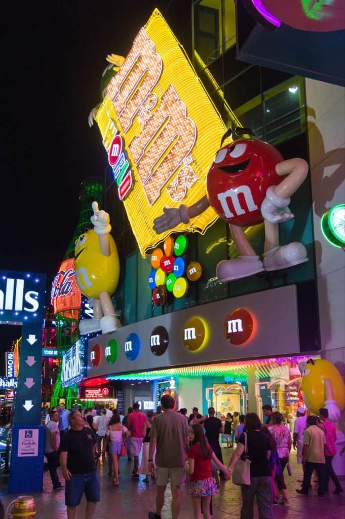 Bright neon M&M's storefront display with colorful character mascots at night in a busy shopping district.