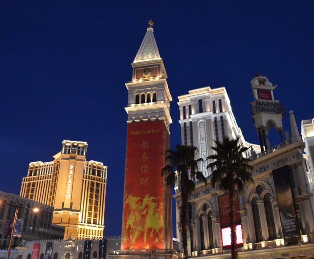 Brightly lit Venetian-style buildings and palm trees at night in Las Vegas.