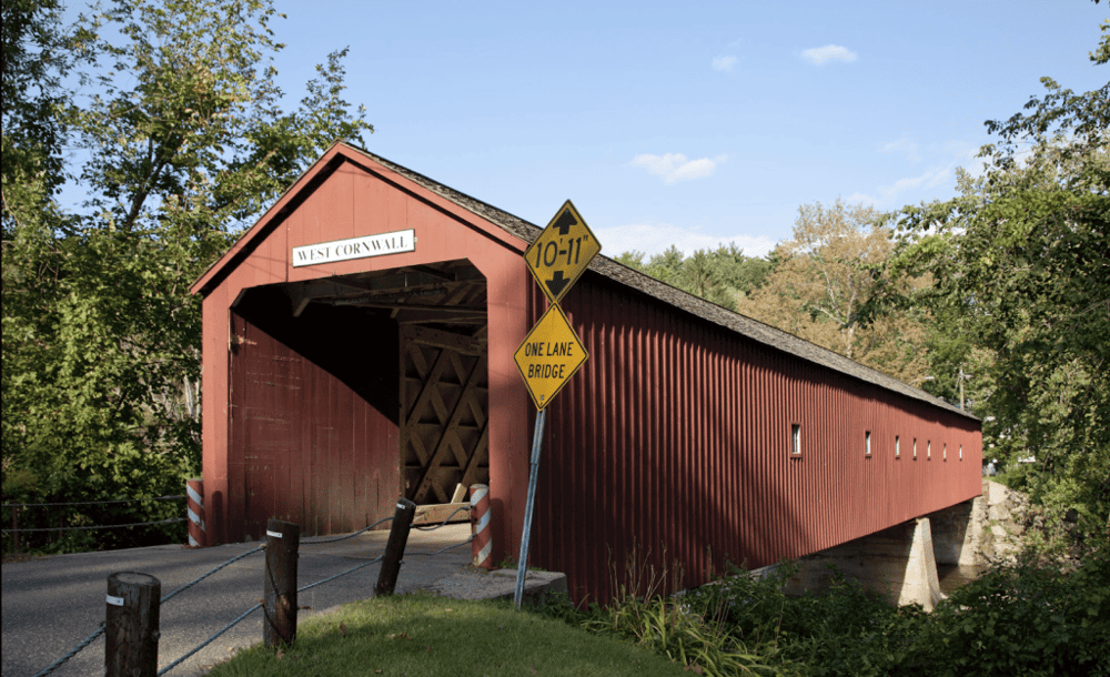 Rustic red covered bridge on a country road with directional signs and lush greenery, ideal for scenic drives and historical exploration.