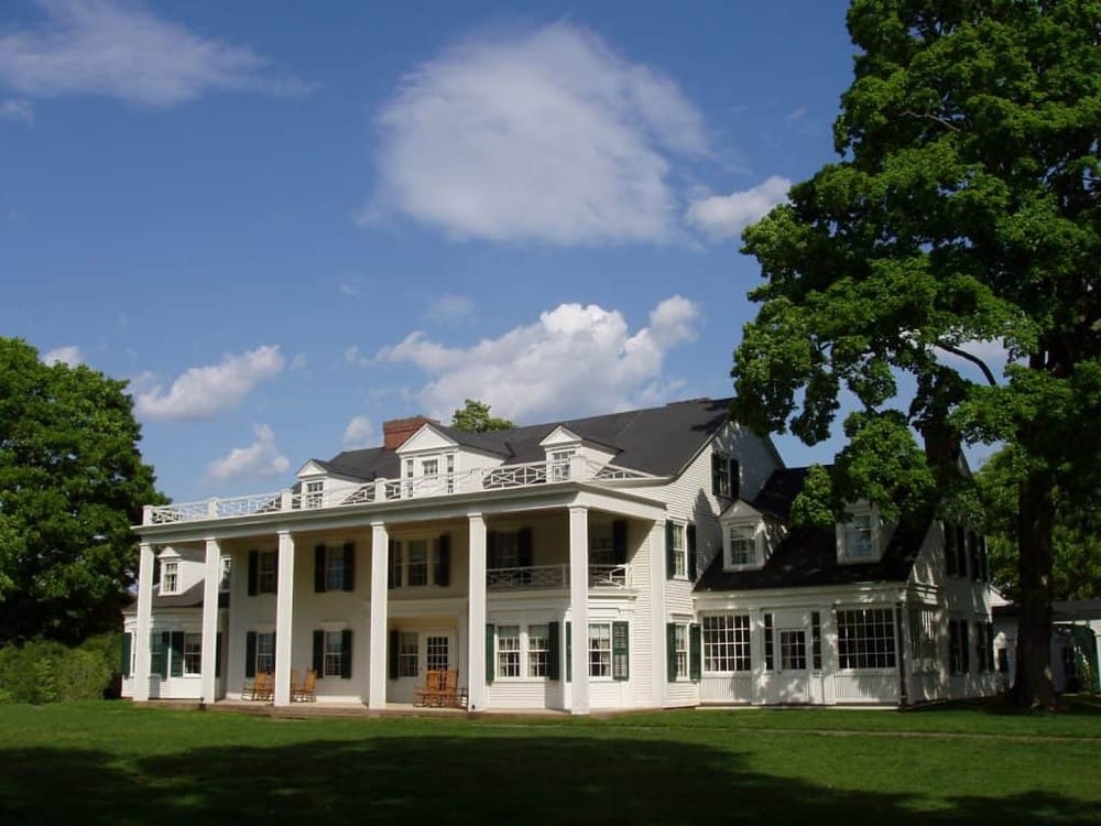 Elegant white mansion with large porch and surrounding green lawn, under a blue sky with clouds.