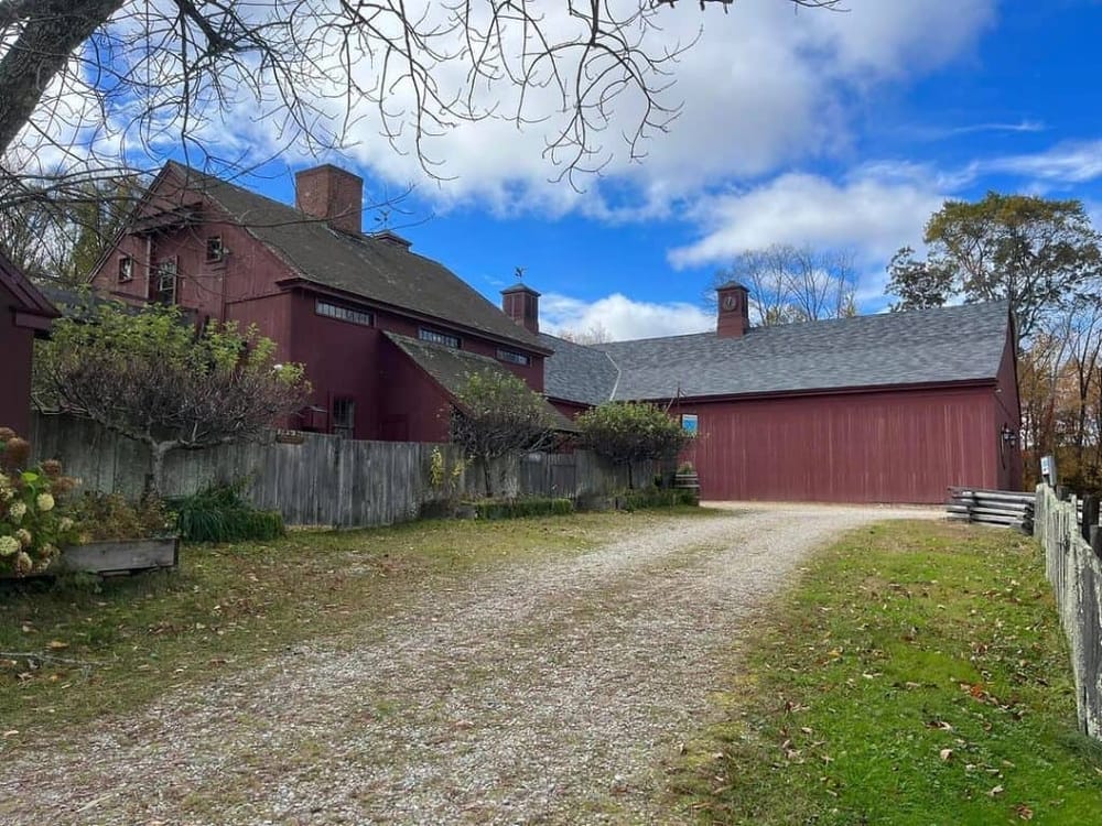 Charming historic red barn with gravel driveway, lush trees, and blue sky, perfect for estate photography or rustic home tours.