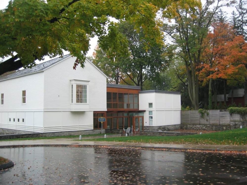Modern house with white siding and large windows, surrounded by autumn trees and wet pavement.