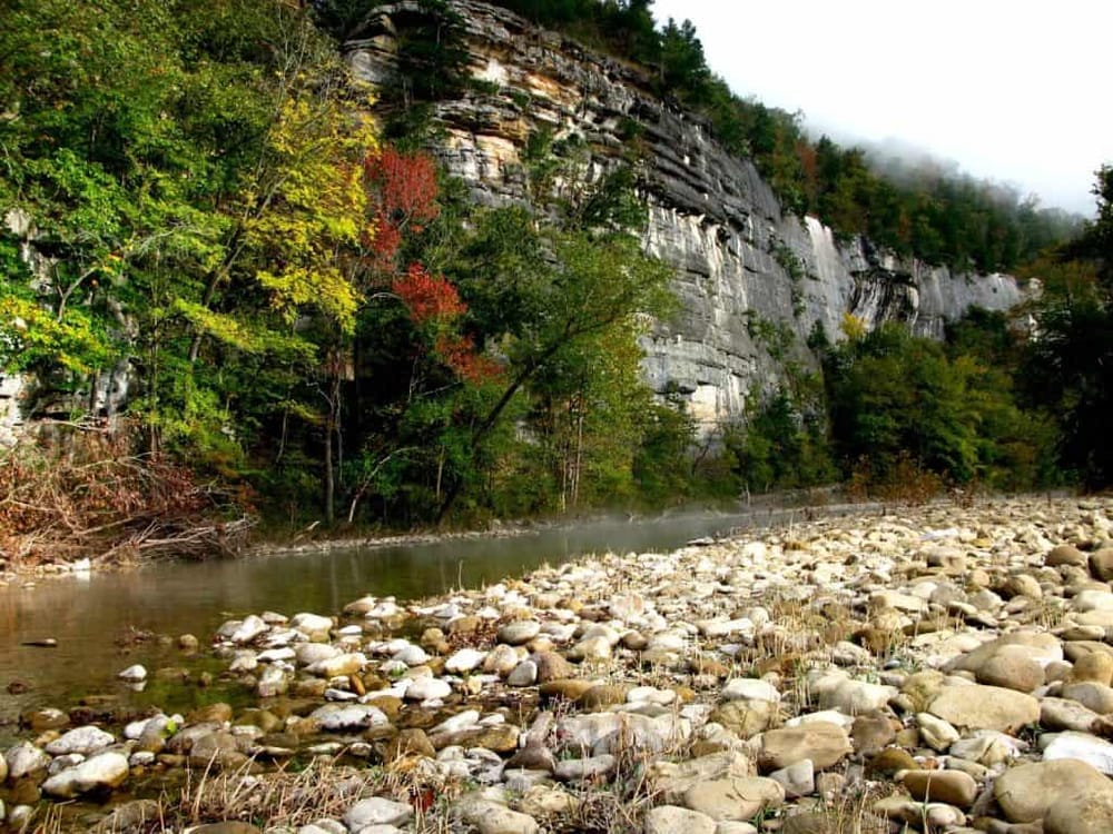 Scenic autumn landscape with rocky riverbed, lush green and colorful trees, and a misty canyon backdrop.