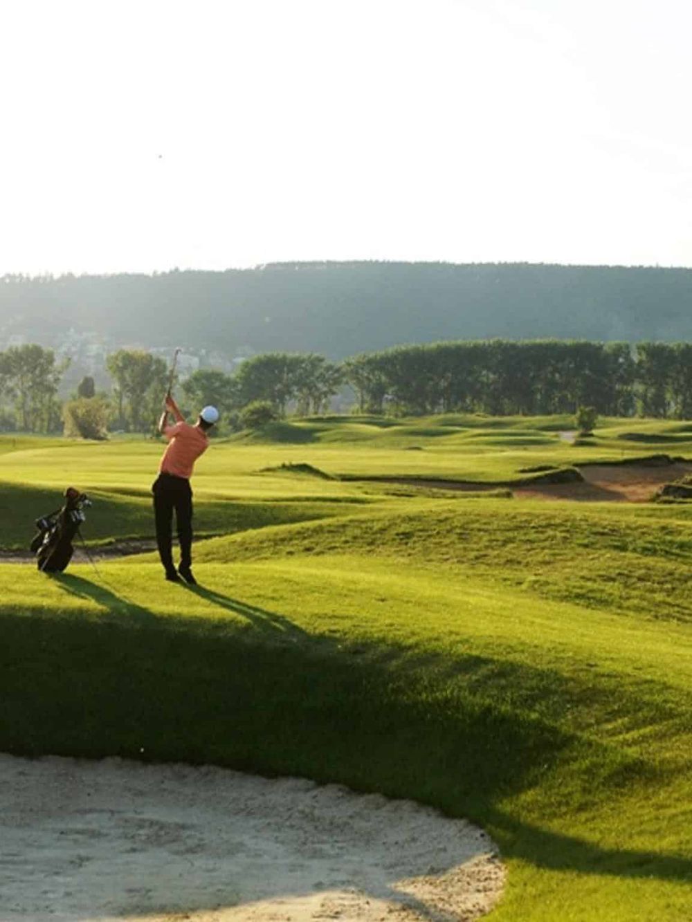 Golf course with a golfer swinging, lush green fairways, and scenic mountain backdrop.