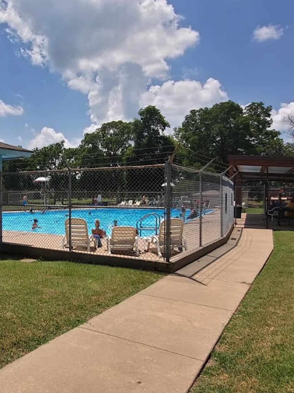 Sunny outdoor swimming pool with people enjoying the water, surrounded by green grass and tall trees.