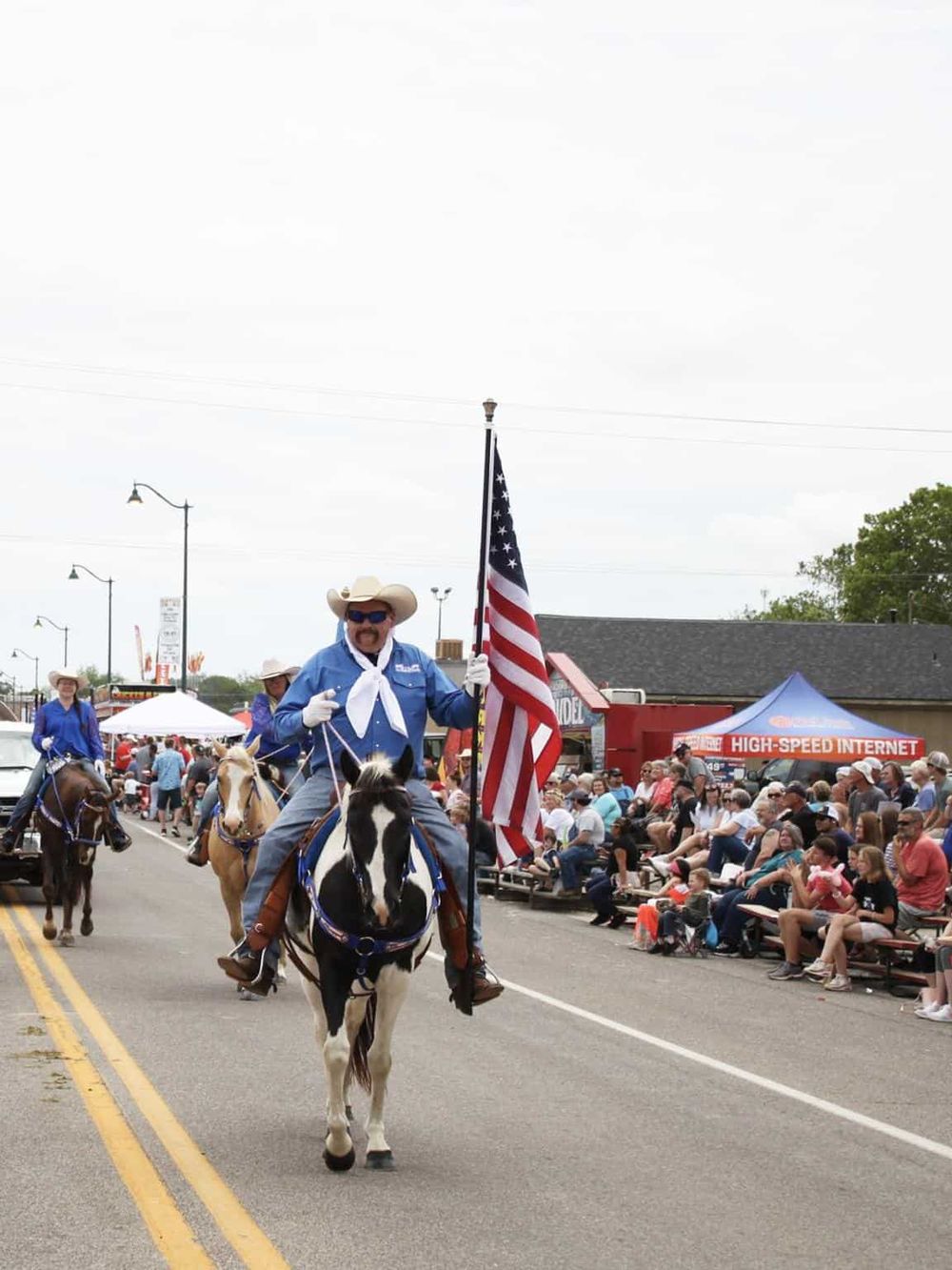 Colorful parade with cowboy on horseback holding American flag, crowds and festival tents in background.