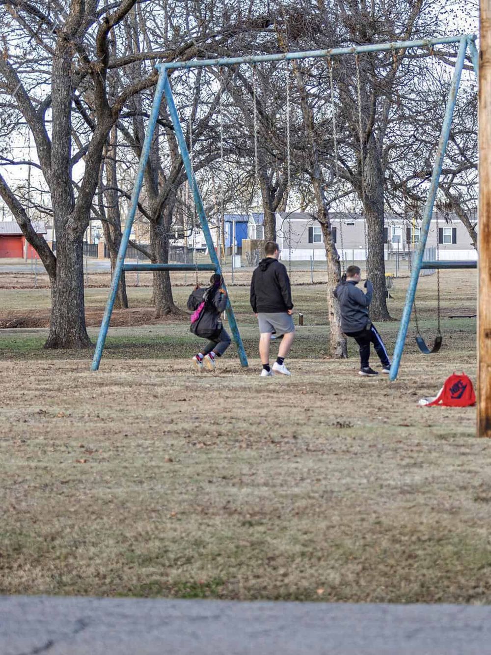 Children playing on swings in an outdoor park with leafless trees and residential buildings in the background.