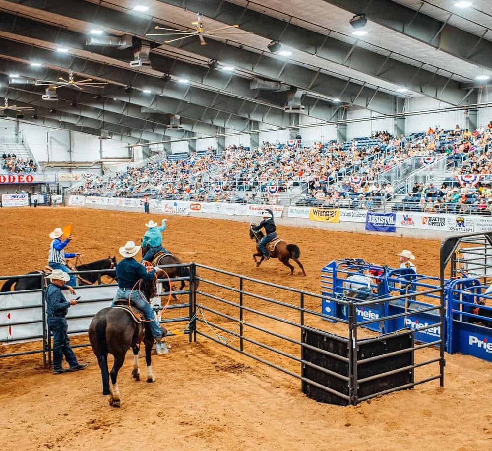 Horse riding competition at indoor arena with spectators, rodeo event, and rodeo signage.