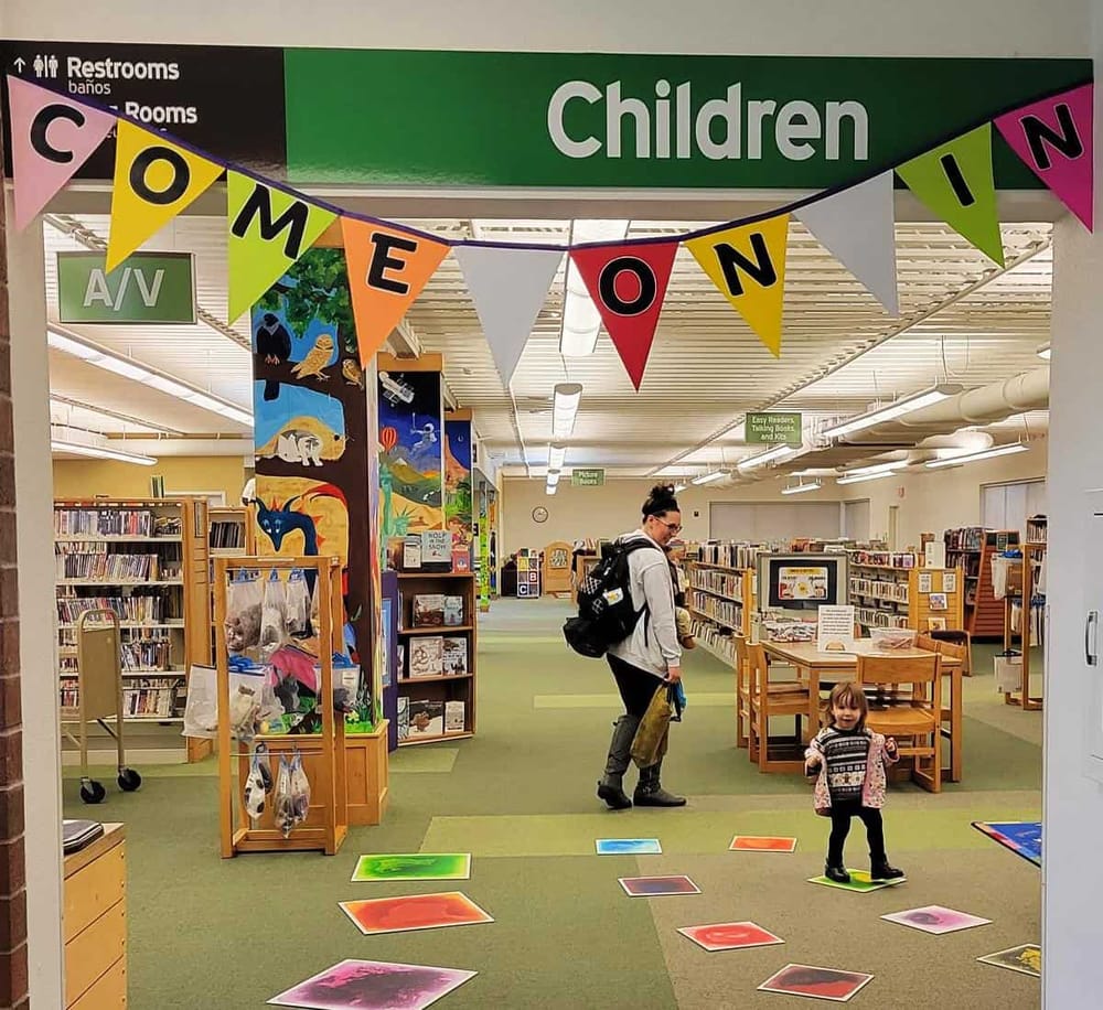 Colorful children's section in public library with signage, artwork, and kids playing on floor mats.