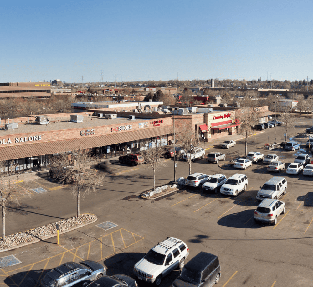Shopping plaza with retail stores, parking lot, clear sky, and nearby commercial buildings.